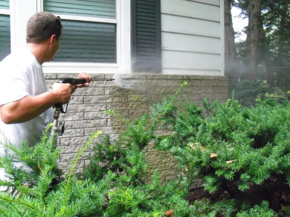 A professional performing exterior cleaning on a brick home using a pressure washer to remove dirt and grime.